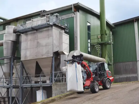 En rød Weidemann Hjullaster 2080 som løfter en stor hvit sekk foran et industribygg.