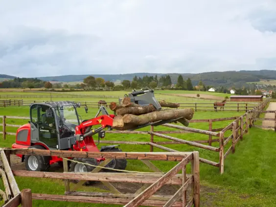 En rød Weidemann Hjullaster 2080 løfter trestokker på en gård med hester i bakgrunnen.