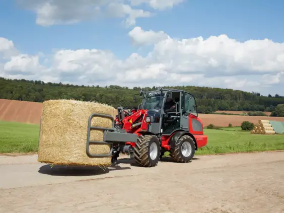 En rød Weidemann Hjullaster 2080 frakter en stor rundballe langs en landevei.