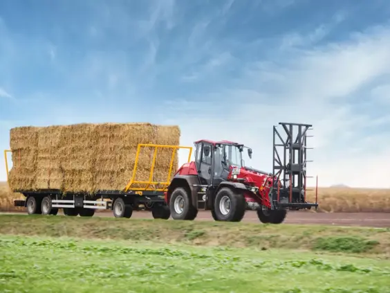 En rød Weidemann 9580T teleskopisk hjullaster trekker en tilhenger med halmballer på et jorde.