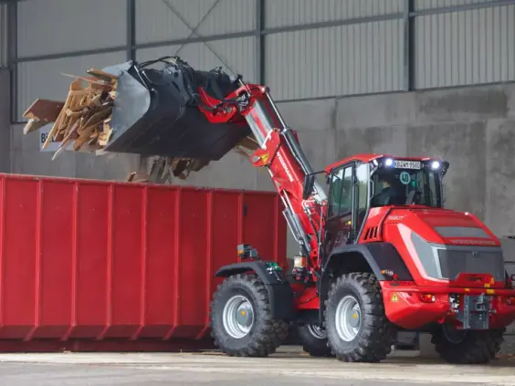 En rød Weidemann 9580T teleskopisk hjullaster som tømmer treavfall i en stor rød container.