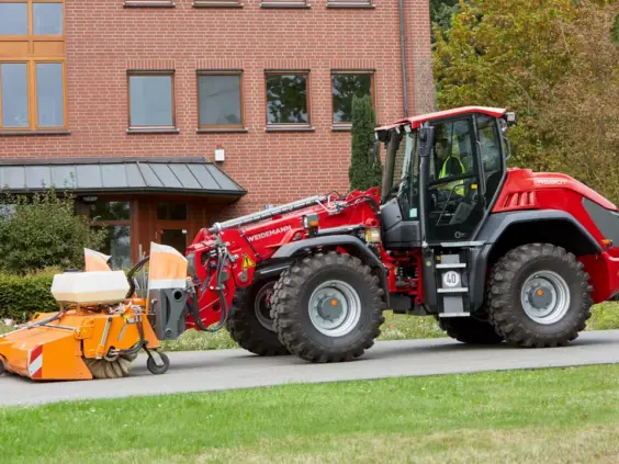 Rød Weidemann 9580T teleskopisk hjullaster utstyrt med en oransje feiemaskin på en vei.