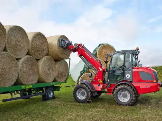 En rød Weidemann 5080T teleskopisk hjullaster som stabler runde høyballer på en tilhenger.
