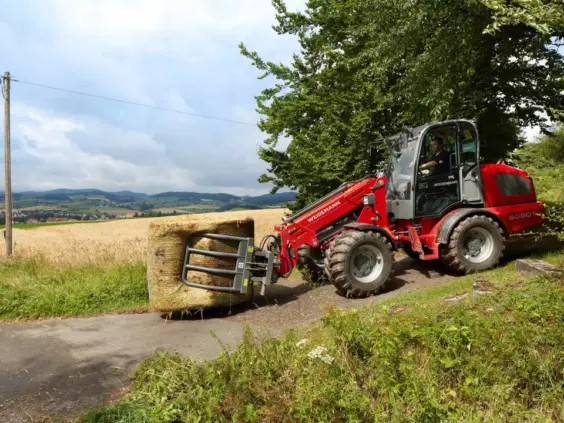Rød Weidemann 5080T teleskopisk hjullaster som transporterer en rundballe på en landevei.