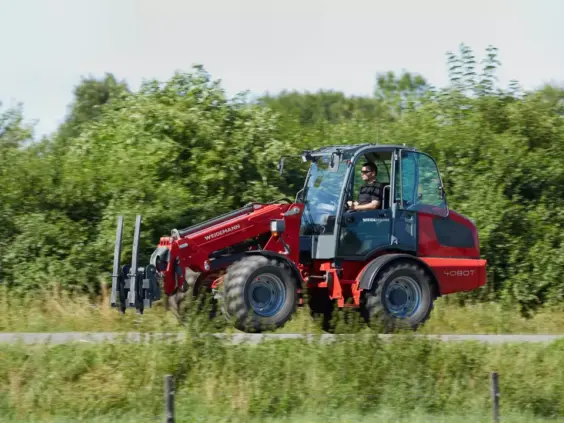 En rød Weidemann 4080T teleskopisk hjullaster i bevegelse på en vei langs grønne trær.