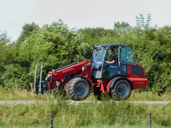 En rød Weidemann 4080T teleskopisk hjullaster i bevegelse på en vei langs grønne trær.