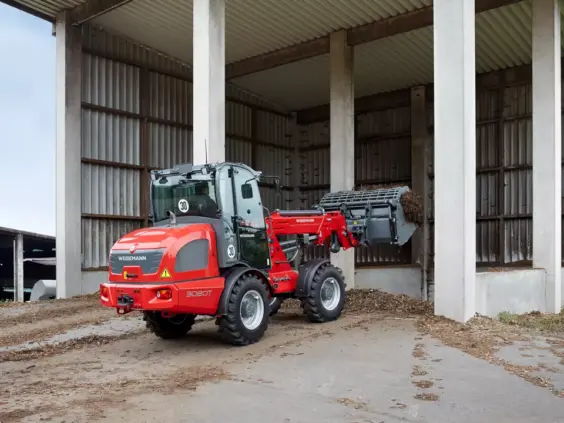 En rød Weidemann 3080T teleskopisk hjullaster i arbeid utenfor et gårdsbygg.