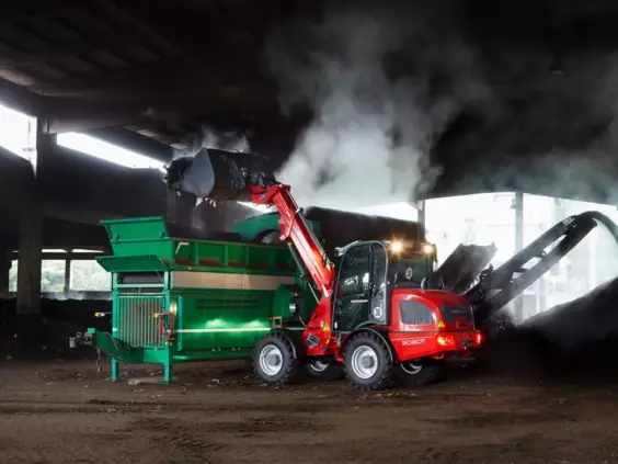 En rød Weidemann 3080T teleskopisk hjullaster som tømmer dampende materiale i en grønn maskin.