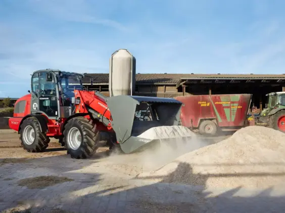 Rød Weidemann 3080T teleskopisk hjullaster som laster tørt materiale på en gård.