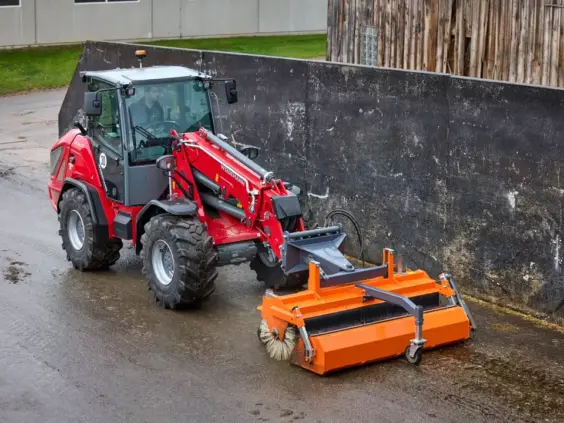 Rød Weidemann 3060T teleskopisk hjullaster med en oransje feiemaskin i arbeid utendørs.