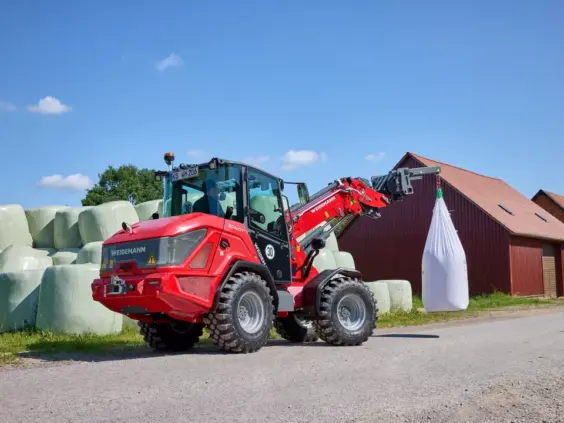 En rød Weidemann 2060T teleskopisk hjullaster løfter en stor hvit sekk på et gårdsbruk.