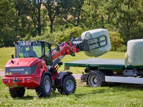 En rød Weidemann 2060T teleskopisk hjullaster som løfter en rundballe opp på en tilhenger.