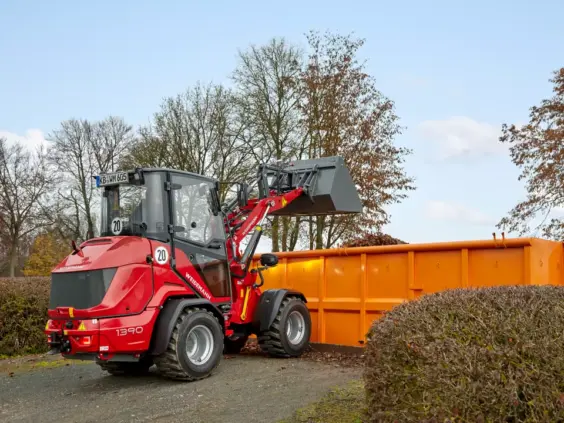 En rød Weidemann Hoftrac 1390 laster avfall i en stor oransje container utendørs.