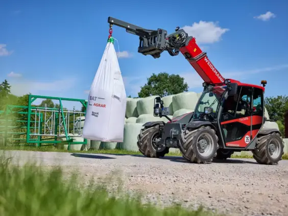 En rød Weidemann teleskoplaster T6025 som løfter en stor hvit sekk utendørs.