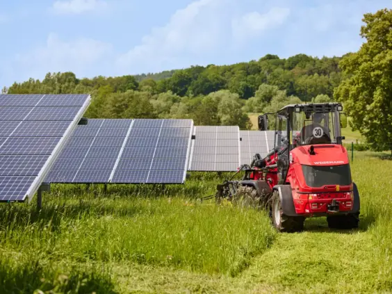 Rød Weidemann Hoftrac 1390 i arbeid med gressklipping ved siden av store solcellepaneler.