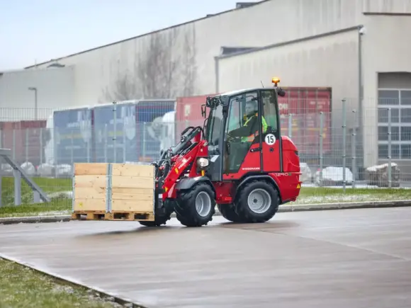 Rød Weidemann Hoftrac 1290e elektrisk minilaster som frakter en trekasse på et industriområde.