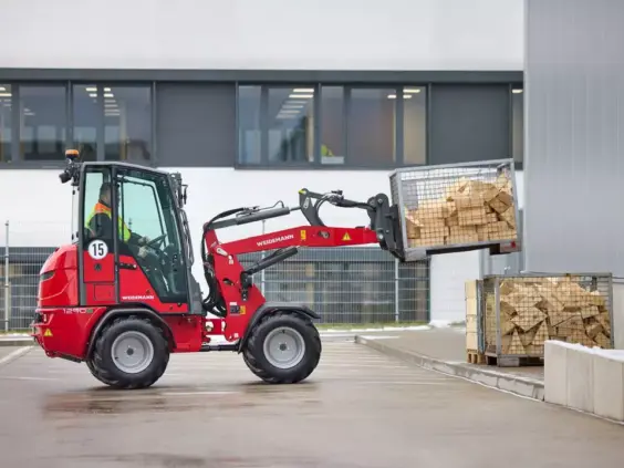 Rød Weidemann Hoftrac 1290e elektrisk minilaster løfter en kasse med treklosser utendørs.