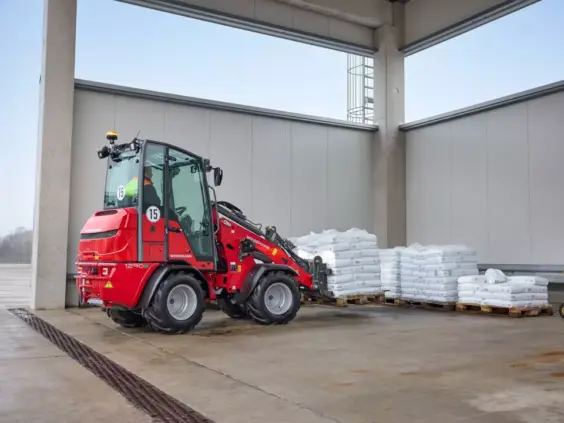 Rød Weidemann Hoftrac 1290e elektrisk minilaster som løfter paller med sekker i et lager.