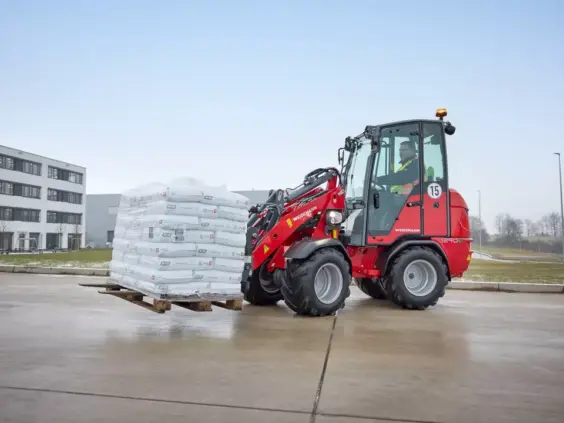 En rød Weidemann Hoftrac 1290e elektrisk minilaster som løfter en pall med sekker.