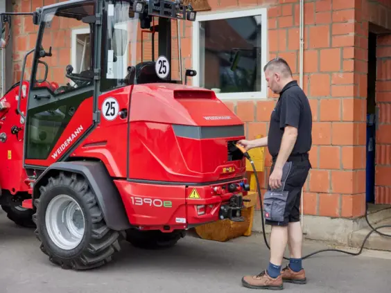 En mann som lader en rød Weidemann Hoftrac 1390e elektrisk hjullaster utendørs.