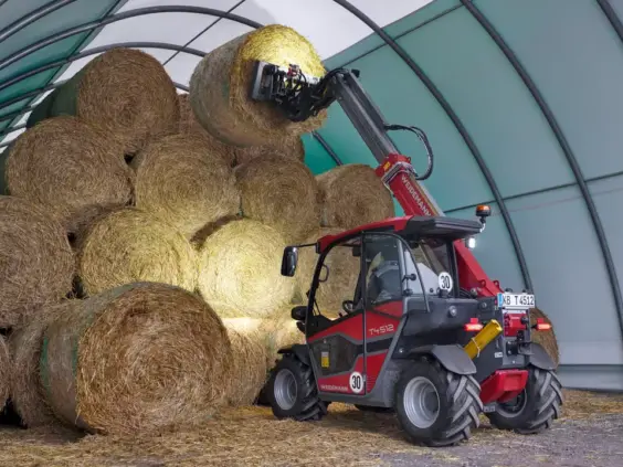 En rød Weidemann teleskoplaster T4512 løfter en rundballe i en tunnelhall.