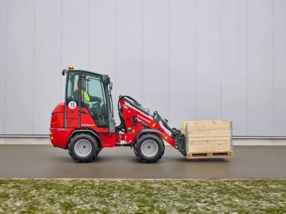 Rød Weidemann Hoftrac 1290e elektrisk minilaster som frakter en trekasse foran en grå vegg.
