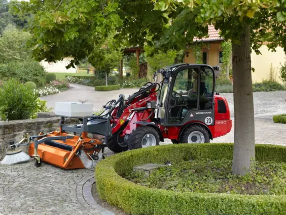 Weidemann Hoftrac 1280 minilaster med påmontert feiemaskin rengjør en brolagt gangvei utendørs.