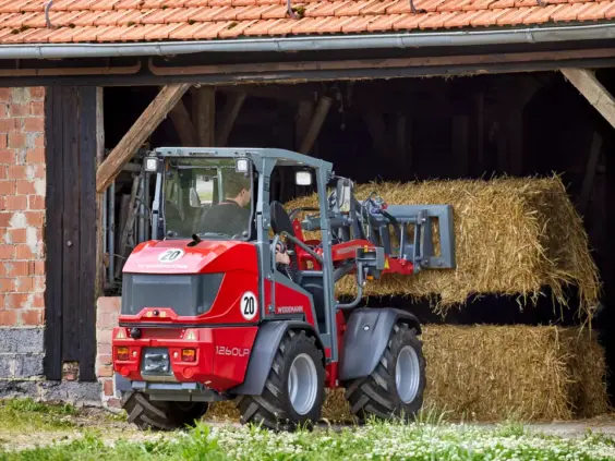Rød Weidemann Hoftrac 1260LP minilaster som løfter en halmballe ved en låve.