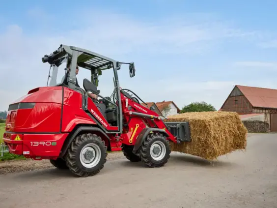 En rød Weidemann Hoftrac 1390e kompaktlaster bærer en stor halmballe på en gårdsvei.