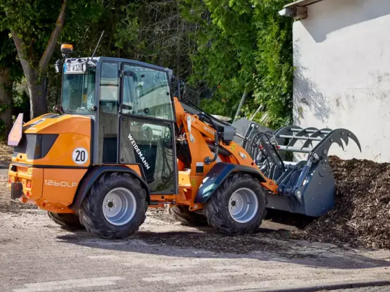 En oransje Weidemann Hoftrac 1260LP minilaster parkert ved en haug med flis utendørs.