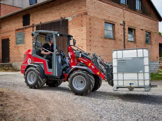 En rød og grå Weidemann Hoftrac 1260LP minilaster transporterer en hvit vanntank på grus.