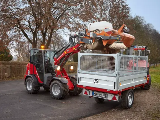 En rød Weidemann Hoftrac 1260LP minilaster tømmer en feiemaskin i en tilhenger.