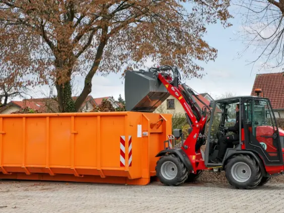 En rød Weidemann Hoftrac 1260LP minilaster tømmer avfall i en stor oransje container.