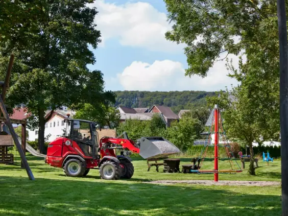 En rød Weidemann Hoftrac 1390e hjullaster fylt med grus på en grønn lekeplass.