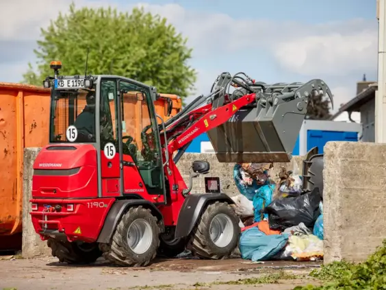 En rød Weidemann Hoftrac 1190e minilaster som tømmer avfall i en container utendørs.