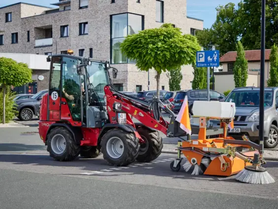 En rød Weidemann Hoftrac 1190e minilaster med påmontert feiemaskin på en parkeringsplass.