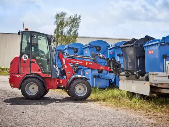 En rød Weidemann Hoftrac 1190e minilaster løfter en avfallsbeholder opp på en tilhenger.