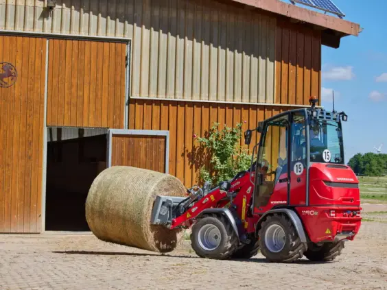 En rød Weidemann Hoftrac 1190e minilaster som flytter en stor rundballe utenfor en låve.