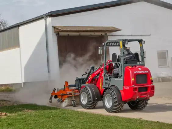 En rød Weidemann Hoftrac 1160 minilaster utstyrt med feiemaskin feier en støvete gårdsplass.