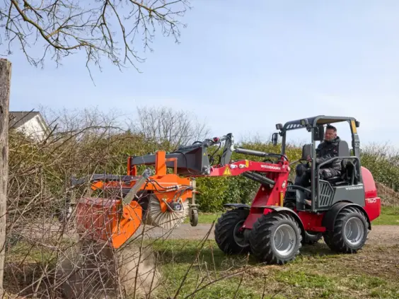 En rød Weidemann Hoftrac 1160 minilaster som fjerner ugress og kvister utendørs.