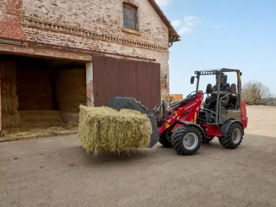 En rød Weidemann Hoftrac 1160 minilaster løfter en stor høyball utenfor en gammel låve.