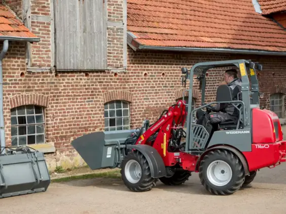 En rød og grå Weidemann Hoftrac 1160 minilaster foran en gammel murbygning.