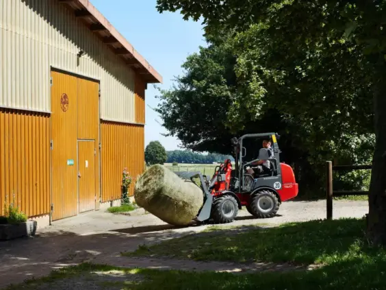 En rød Weidemann Hoftrac 1160 minilaster flytter en rundballe utenfor en låve.