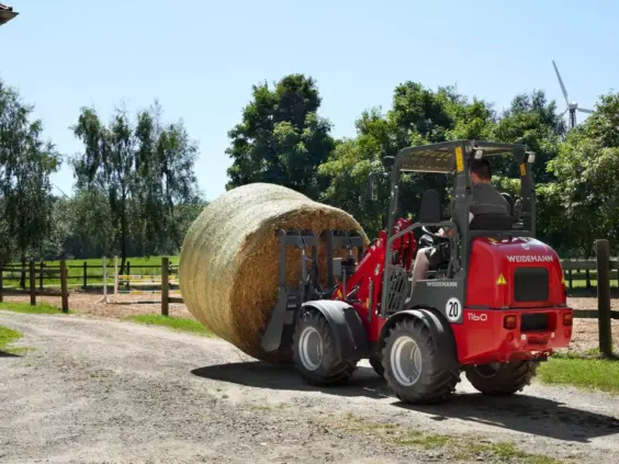 En rød Weidemann Hoftrac 1160 minilaster som transporterer en stor rundballe på en gårdsvei.