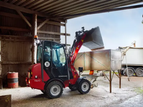 En rød Weidemann Hoftrac 1160 minilaster fyller en beholder på en gård.