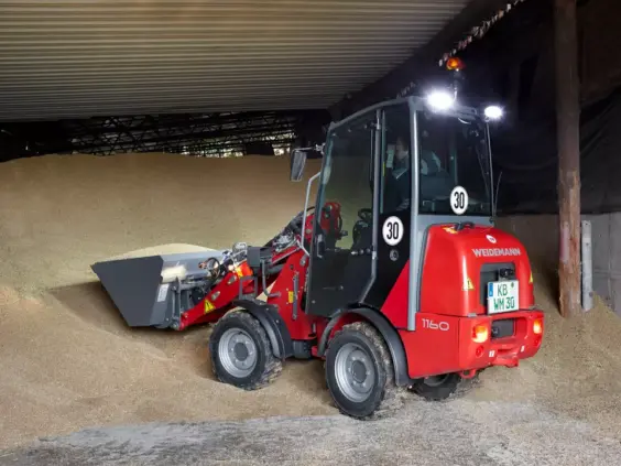 Rød Weidemann Hoftrac 1160 minilaster som flytter korn i en lagerbygning.