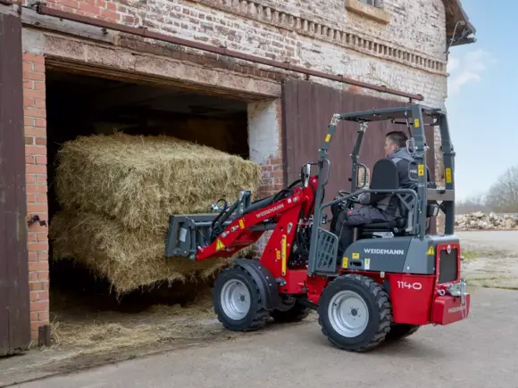 En Weidemann Hoftrac 1140 minilaster løfter en stor høyball ut av en låve.