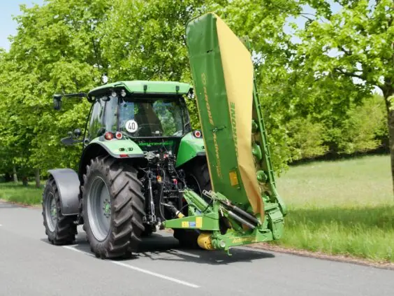 En grønn traktor på veien med en Krone ActiveMow slåmaskin montert bak.