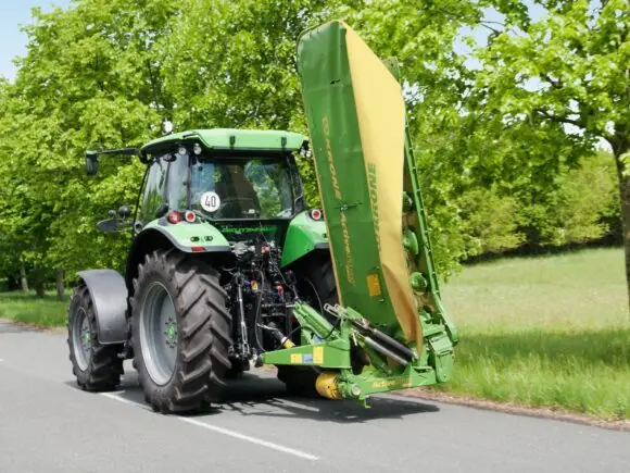En grønn traktor på veien med en Krone ActiveMow slåmaskin montert bak.