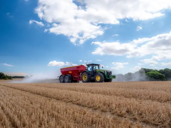 En grønn traktor sprer kalk på en høstet kornåker under en blå himmel.